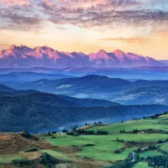 Widok na pasmo górskie o zachodzie słońca, z zielonymi wzgórzami i lasami w tle.